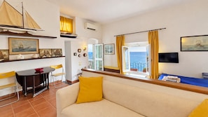A living room with a sofa, a dining table with chairs, a television, and a view of the sea through an open window. - Casa Amanda: A Window onto the Sea in the Portofino Marine Park (Pedale)