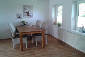 A dining area with a wooden table and wicker chairs, a white radiator, and a window with a view of greenery. - Holiday home with 1 bedroom (Langballig)