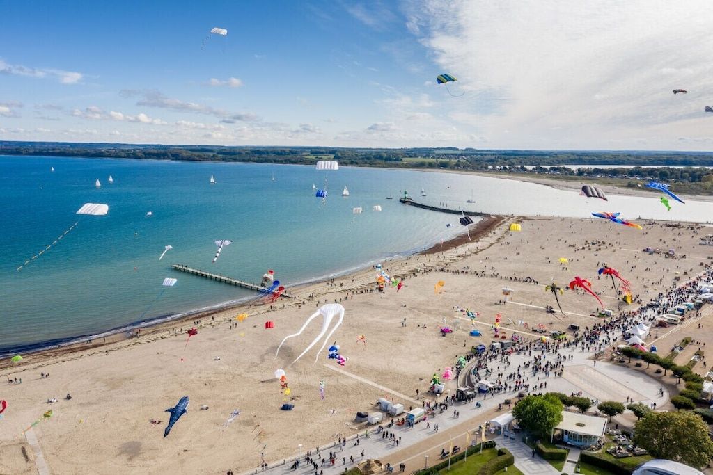 Une plage avec une jetée qui s’avance dans l’eau, entourée d’une foule de personnes et de nombreux cerfs-volants.