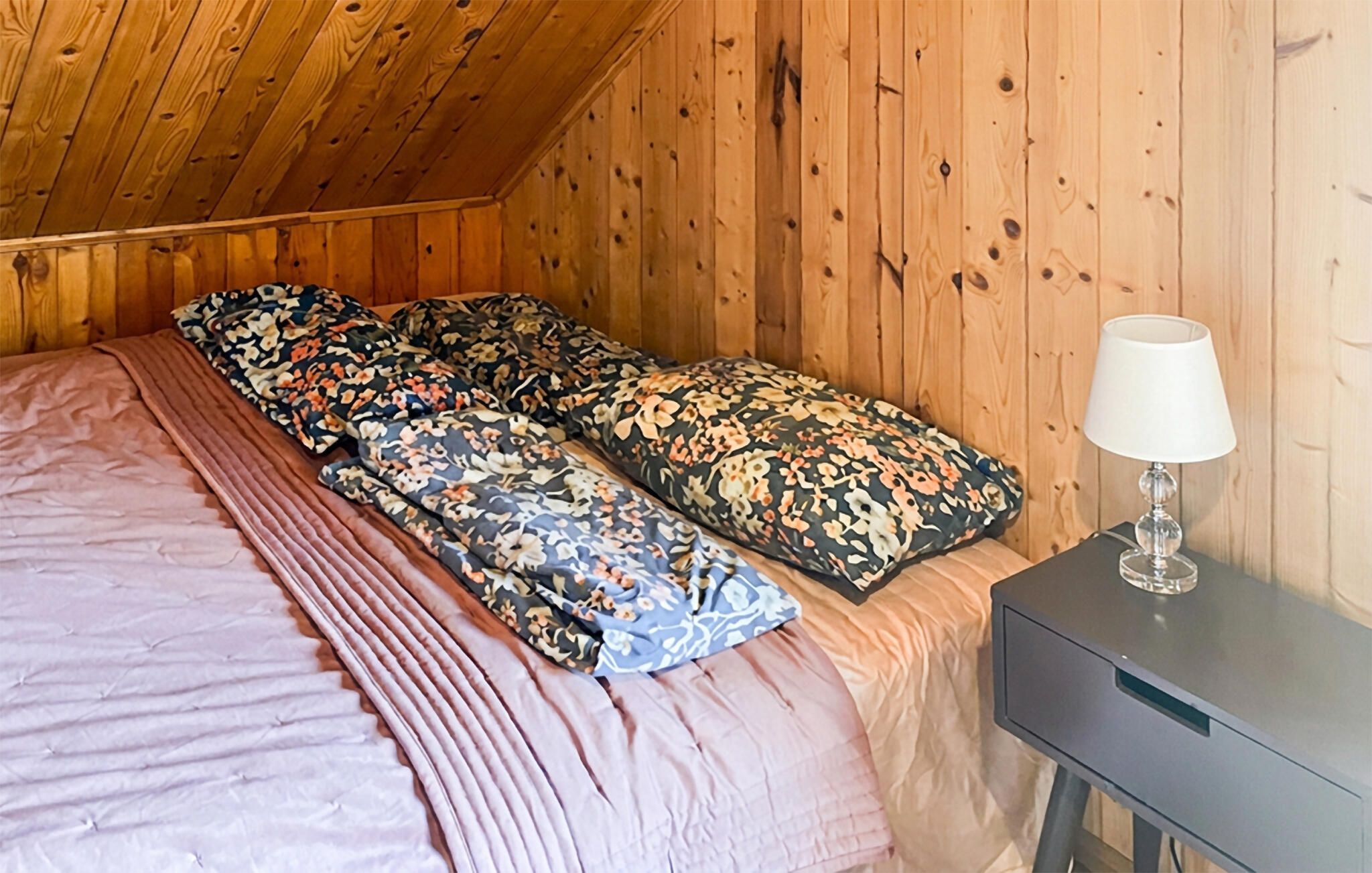 Quarto com teto de madeira, cama com roupa de cama florida e mesa de cabeceira com abajur.