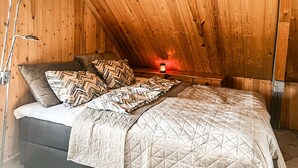 Un dormitorio con techo inclinado de madera, una cama con colcha acolchada, varios almohadones y una mesita de noche con lámpara.