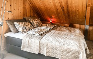 A bedroom with a wooden sloped ceiling, a bed with a quilted bedspread, multiple pillows, and a bedside table with a lamp.
