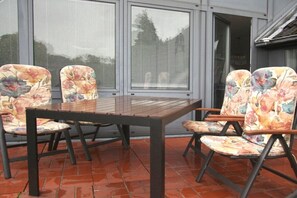 Eine Außenpatio mit einem Holztisch und Stühlen, großen Fenstern mit Jalousien und einem gefliesten Boden.