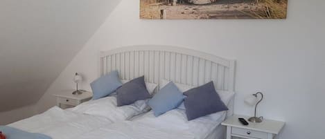 A bedroom with a bed, pillows, a bedside table with a lamp, and a wall-mounted picture of a beach.