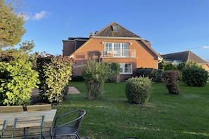 A two-story house with a brick exterior, a balcony, and a well-maintained garden with various shrubs and a wooden bench. - Holiday apartment with 1 bedroom (Boltenhagen)
