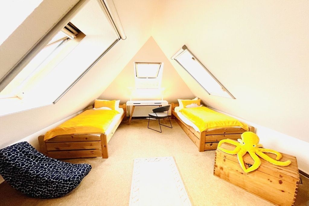 Un dormitorio bajo cubiertas, pequeño y triangular, con dos camas, un escritorio y una silla.