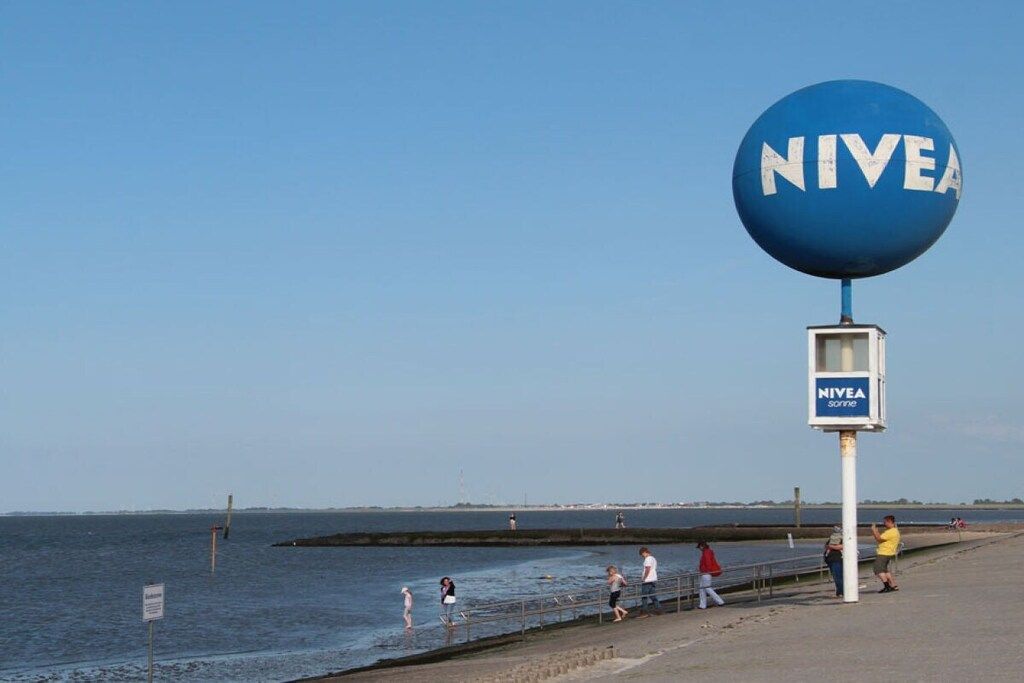 Una spiaggia con un grande cartello blu con scritto 'NIVEA' e un cartello più piccolo sotto.