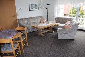 A room with a grey sofa, a wooden table, and a chair. There is a window with a view of buildings and trees. - 3 ( OR) with balcony "House Sloop" (Dornumersiel)