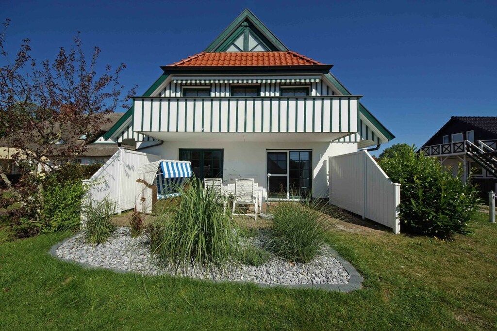 Une maison à deux étages, avec un toit recouvert de tuiles rouges, des murs extérieurs blancs et un petit porche à l’avant, agrémenté de chaises.