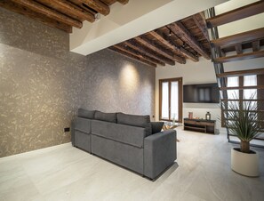 A modern living room with a grey sofa, wooden ceiling beams, a flat-screen TV, and a large window with natural light. - Cà Delle Navi Apartment R&R (Venice)