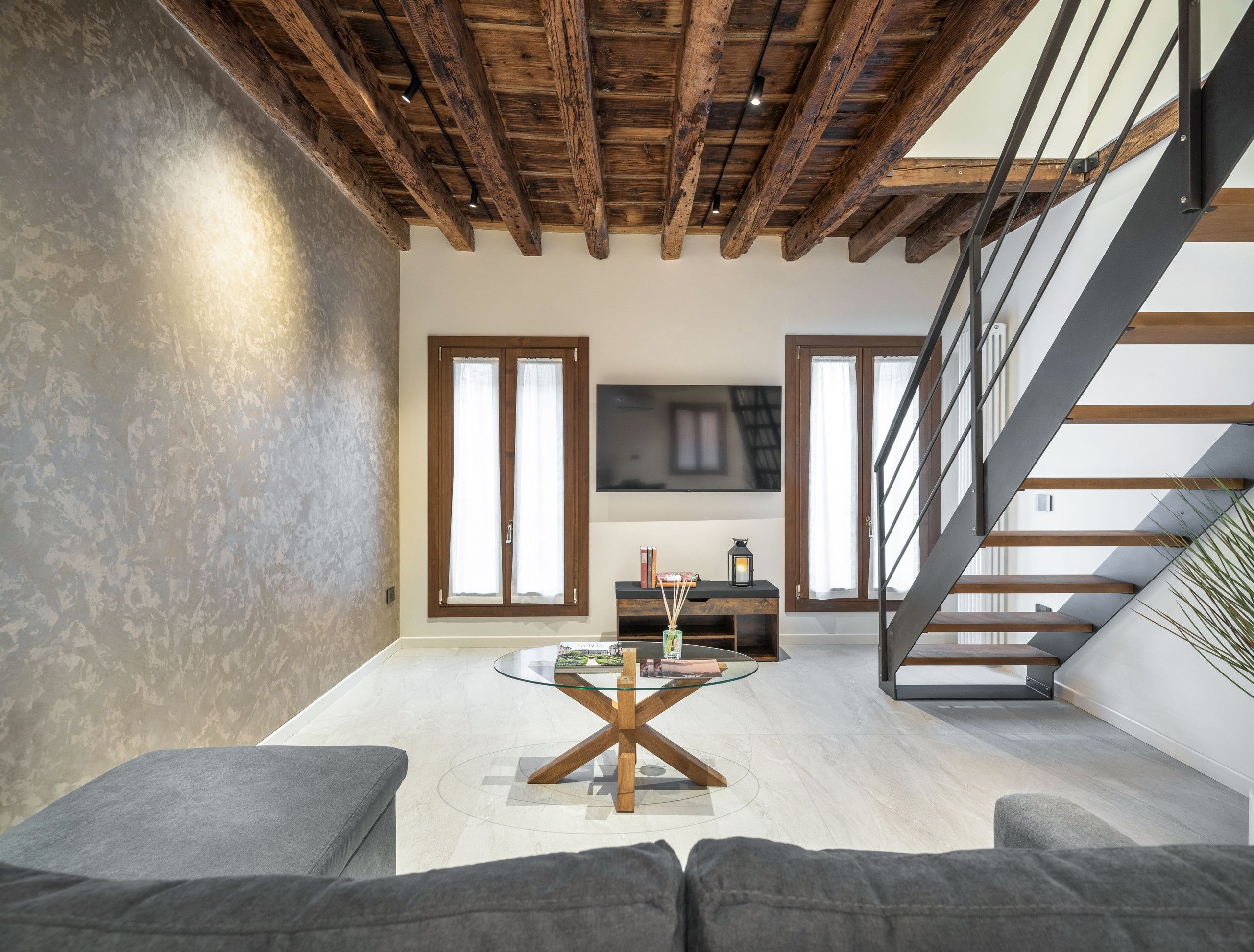 Un salon moderne avec une table basse en verre, des canapés gris, un plafond en bois et un escalier.