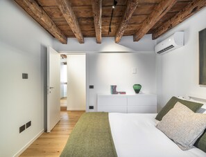 A bedroom with a wooden ceiling, a white bed, a bedside table with books, and an air conditioning unit. - Cà Delle Navi Apartment R&R (Venice)