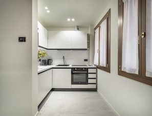 A compact kitchen with white cabinets, a black countertop, a built-in oven, and a window with curtains. - Cà Delle Navi Apartment R&R (Venice)