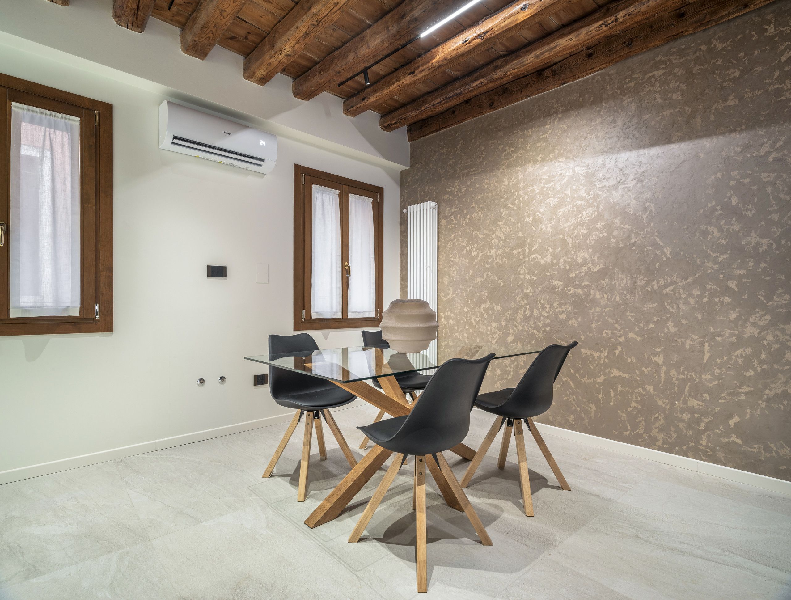 Un comedor moderno con una mesa de vidrio, sillas negras, vigas de madera y una pared con textura.