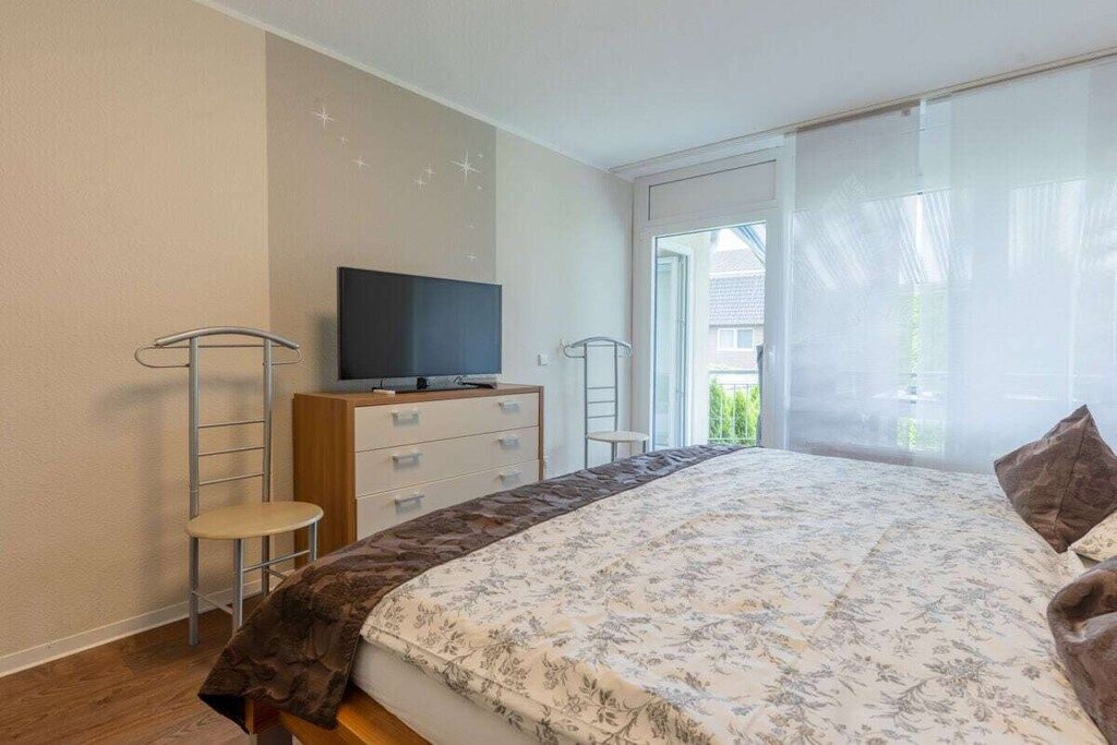 Een slaapkamer met een bed, een televisie op een houten kast, een klein krukje en een raam met uitzicht op groen.