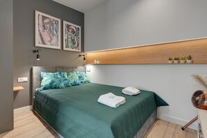 A modern bedroom with a bed, a headboard, wall-mounted artwork, a bedside table, and a small shelf with plants. - Apartament Tamara by Q4 Apartments  (Gdańsk)