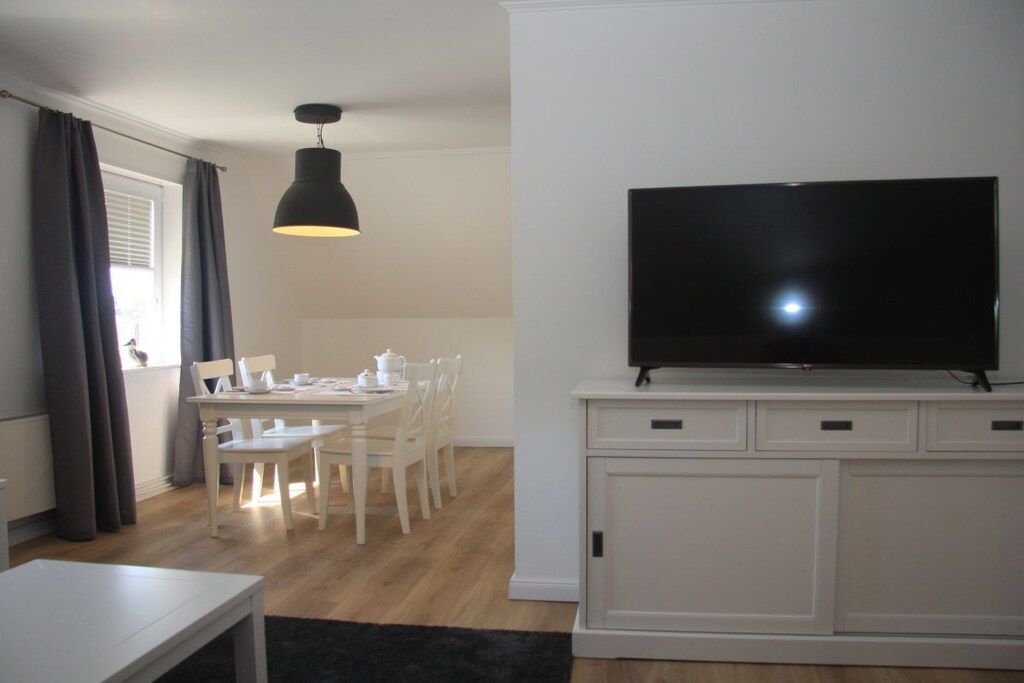 Een moderne woonkamer met een flatscreen televisie op een witte kast, een eettafel met stoelen en een hanglamp.
