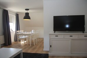 Ein modernes Wohnzimmer mit Flachbildfernseher auf einem weißen Schrank, einem Esstisch mit Stühlen und einer Pendelleuchte.