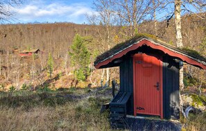 A small wooden cabin with a red door, surrounded by a forested landscape. - 3 bedroom amazing home in Tjørhom (Tjørhom)
