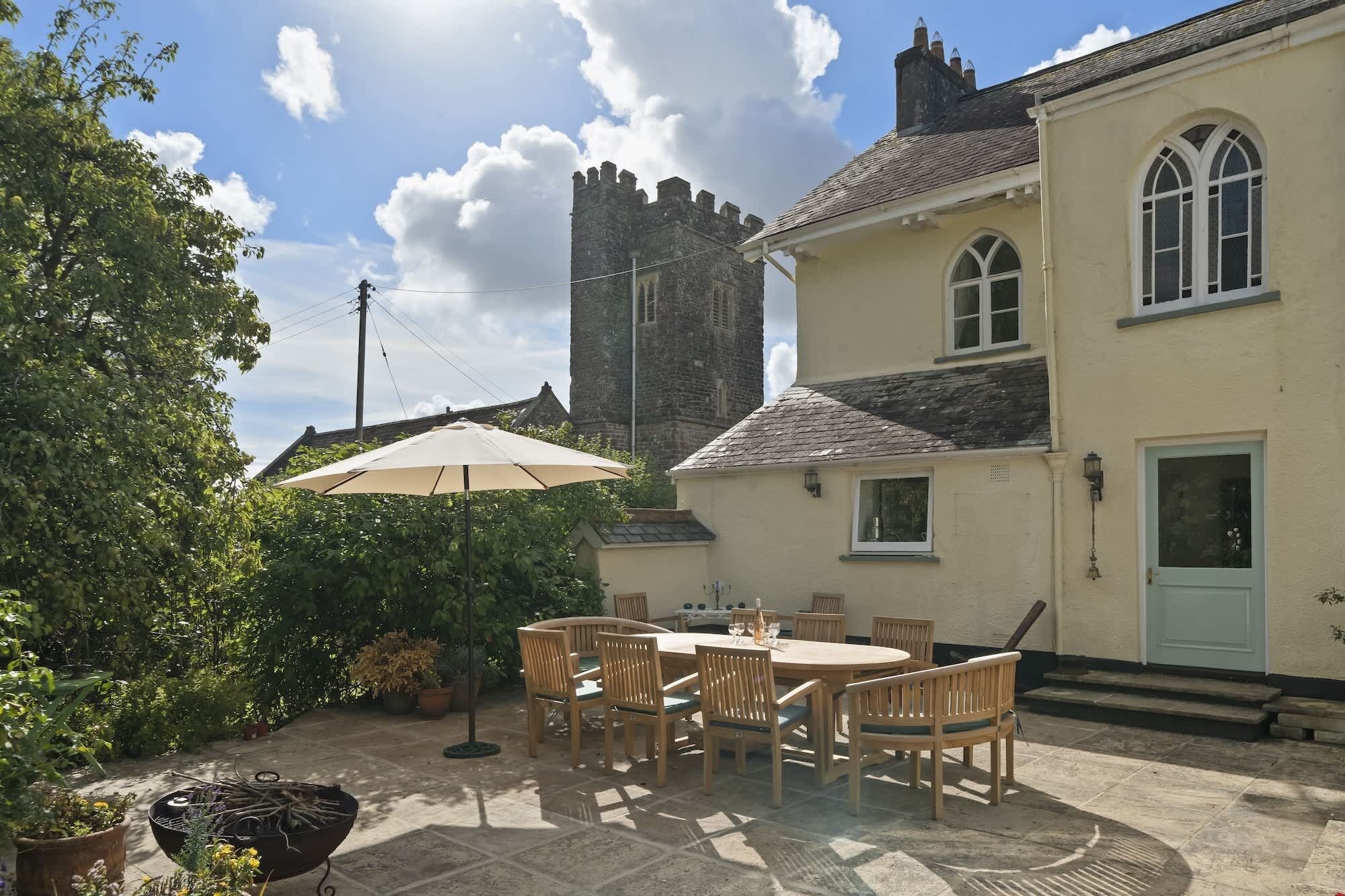 A patio with a table and chairs, a large umbrella, and a stone building with a tower in the background.