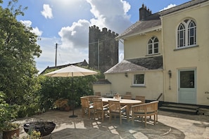 A patio with a table and chairs, a large umbrella, and a stone building with a tower in the background. - Grade II Listed House & Garden in Idyllic Village (Poughill)
