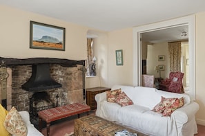 A cozy living room with a stone fireplace, a white sofa with floral cushions, and a bench with a patterned cushion. - Grade II Listed House & Garden in Idyllic Village (Poughill)