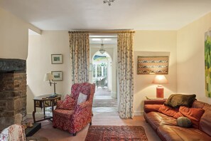 A living room with a fireplace, a red armchair, a brown leather sofa, and a patterned rug. - Grade II Listed House & Garden in Idyllic Village (Poughill)