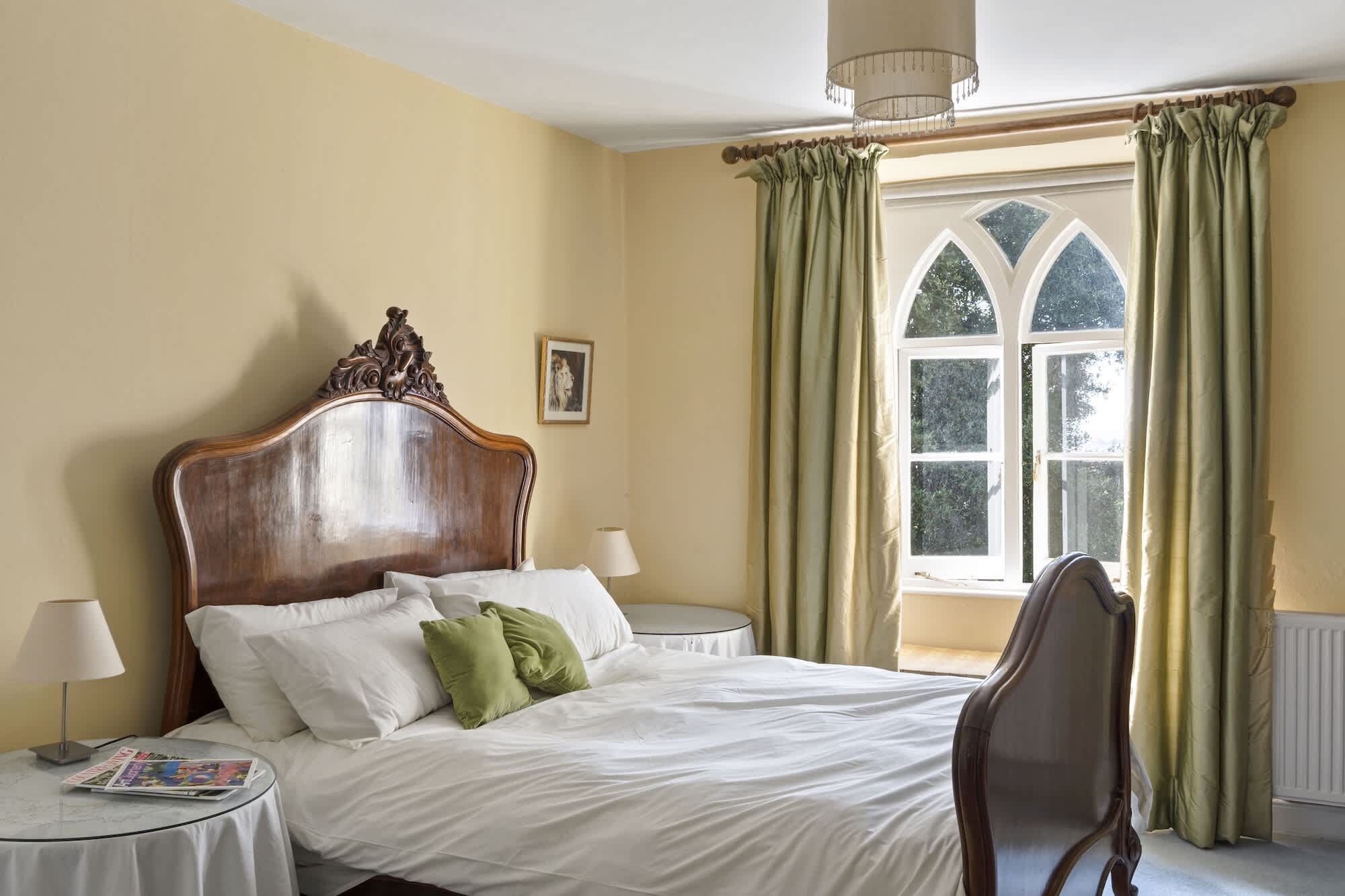 A bedroom with a large bed, a round side table, a chair, a window with curtains, and a chandelier.