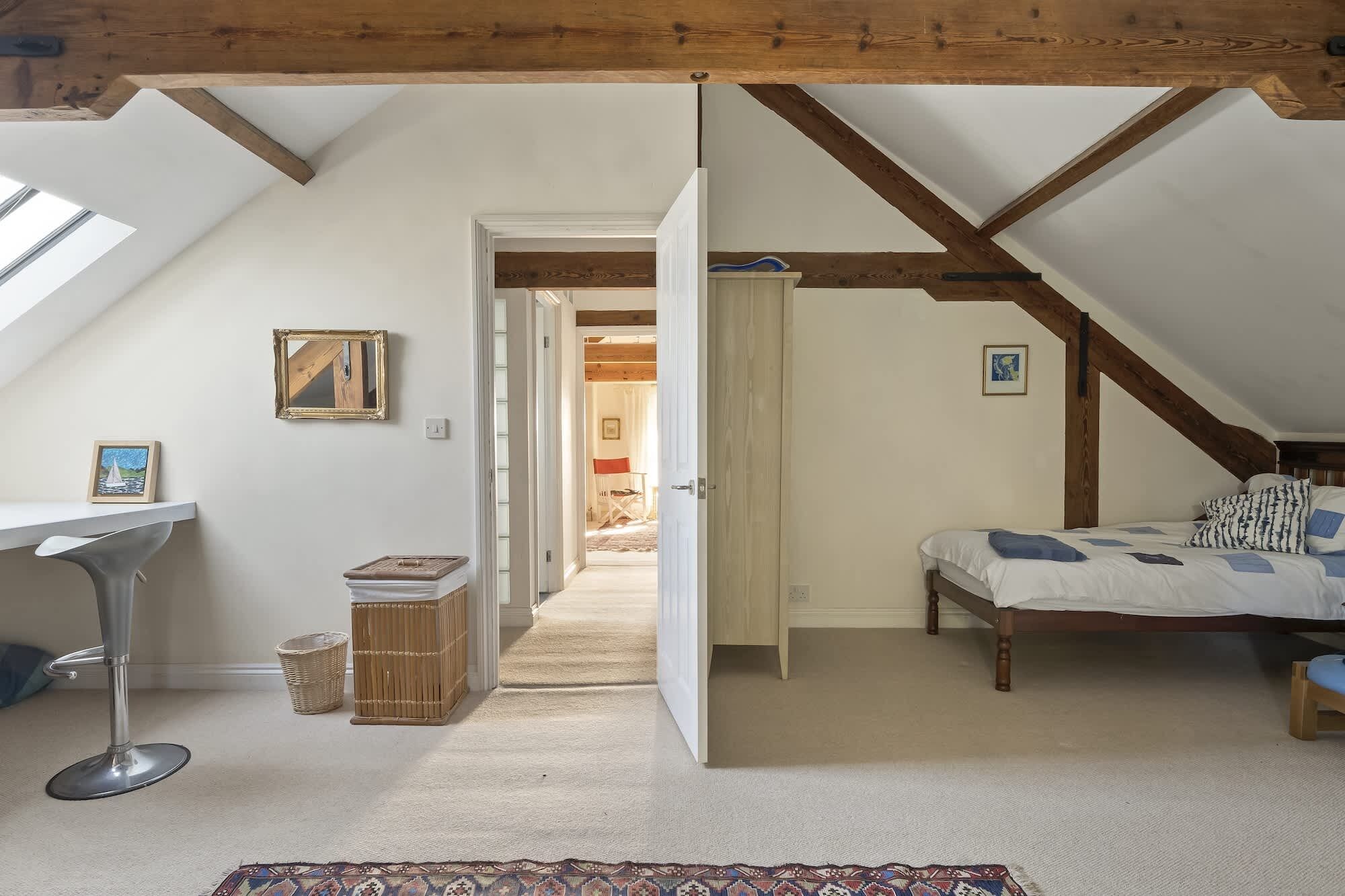 Una habitación con techo inclinado, una cama con cubrecama estampado, una mesita con una silla, y un marco de cuadro en la pared.