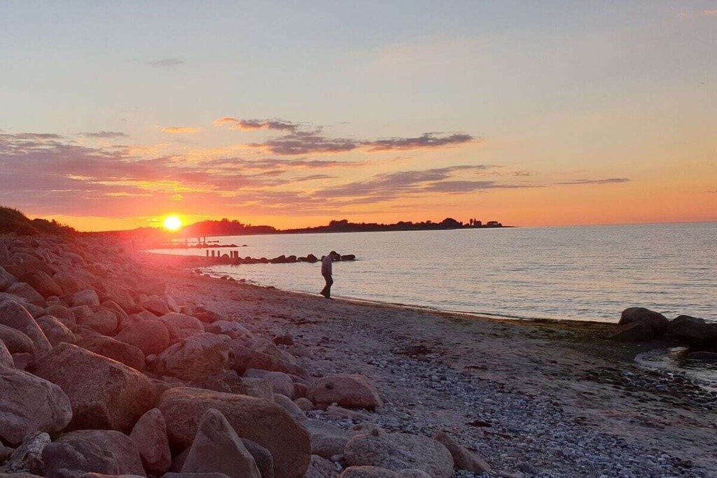 Een strand met grote rotsen, een persoon wandelend en een zonsondergang boven het water.