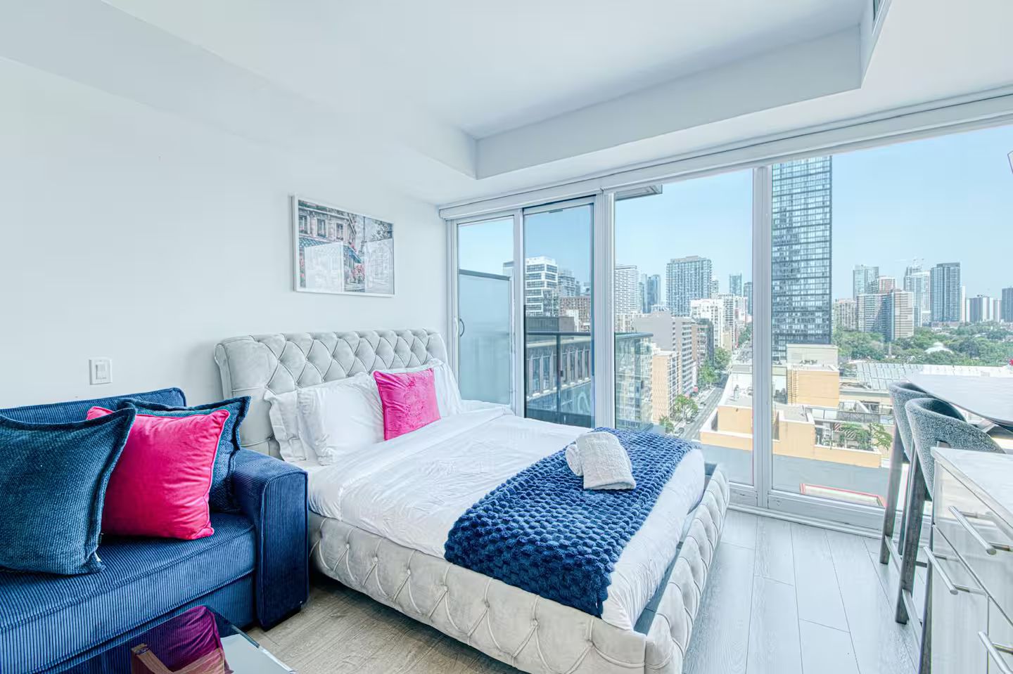 A modern bedroom with a large bed, a sofa, and a city view through a floor-to-ceiling window.