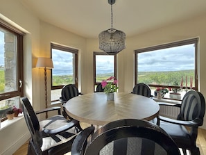 A dining area with a round table, chairs, a hanging light fixture, and large windows offering a view of the landscape. - Cottage 516 - Clifden - sleeps 6 guests  in 3 bedrooms (Clifden)