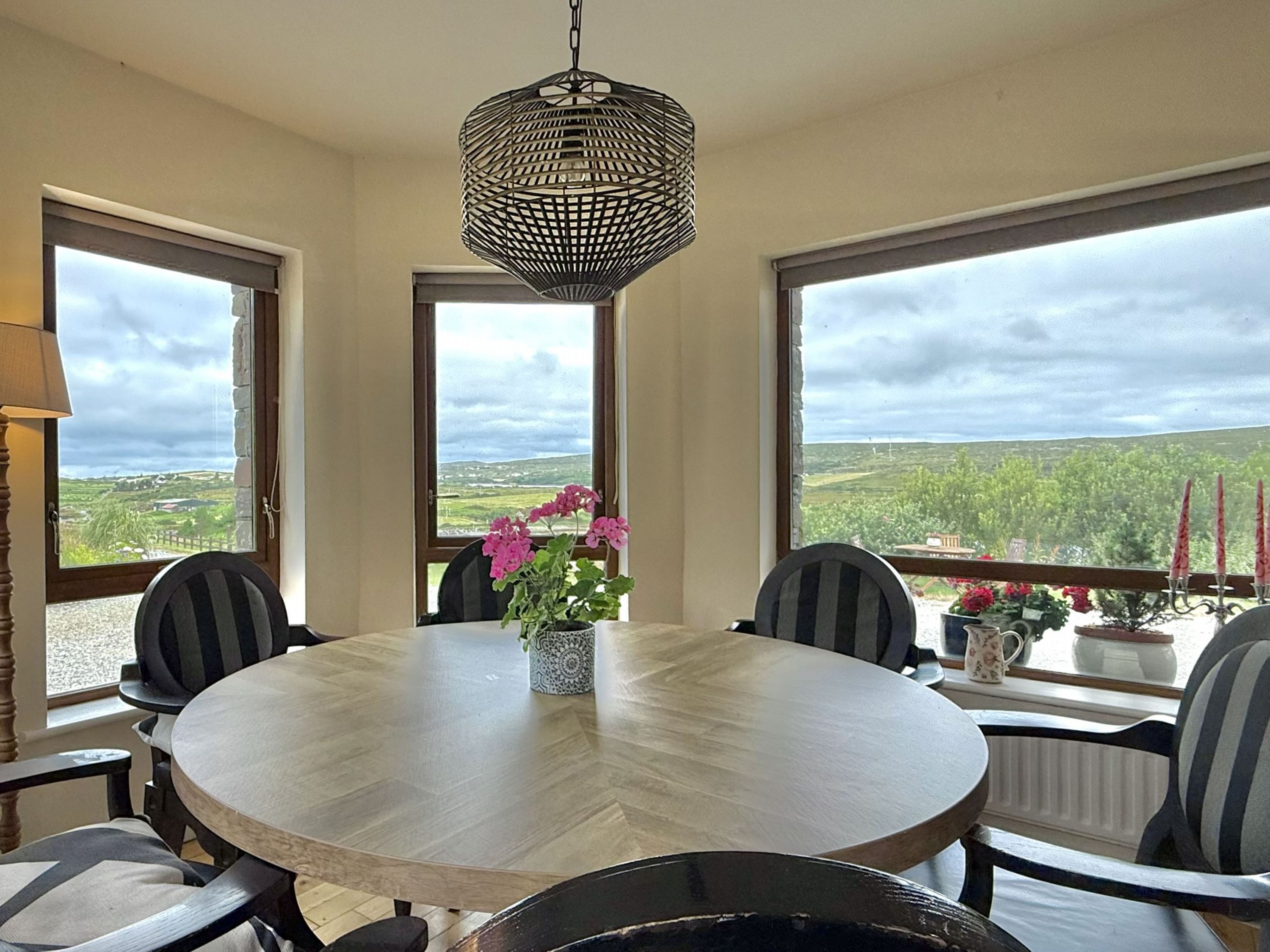 A round dining table with six chairs, a hanging light fixture, and a view of the countryside through large windows.