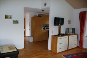 A room with a television on a wooden cabinet, a kitchen area in the background, and a red curtain on the right. - Holiday home with 1 bedroom (Otterndorf)