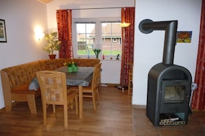 A cozy living room with a sofa, dining table, chairs, a fireplace, and a window with curtains. - Holiday home with 1 bedroom (Otterndorf)