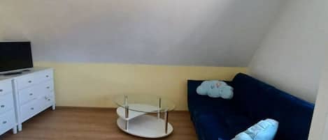 Une pièce avec une commode blanche, une table en verre, un canapé bleu et un téléviseur posé sur la commode.
