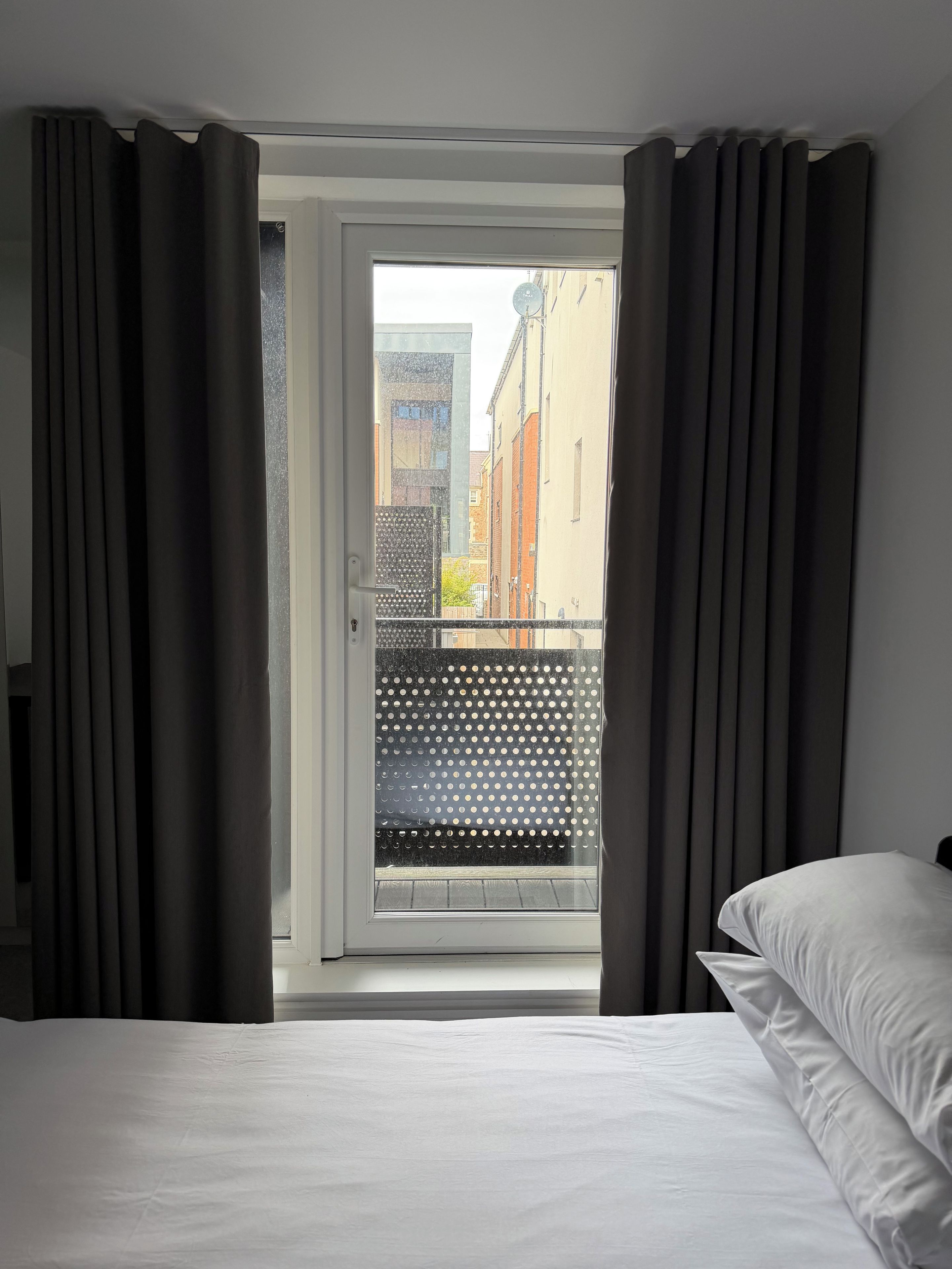 A hotel room with a bed, a window with curtains, and a view of buildings outside.