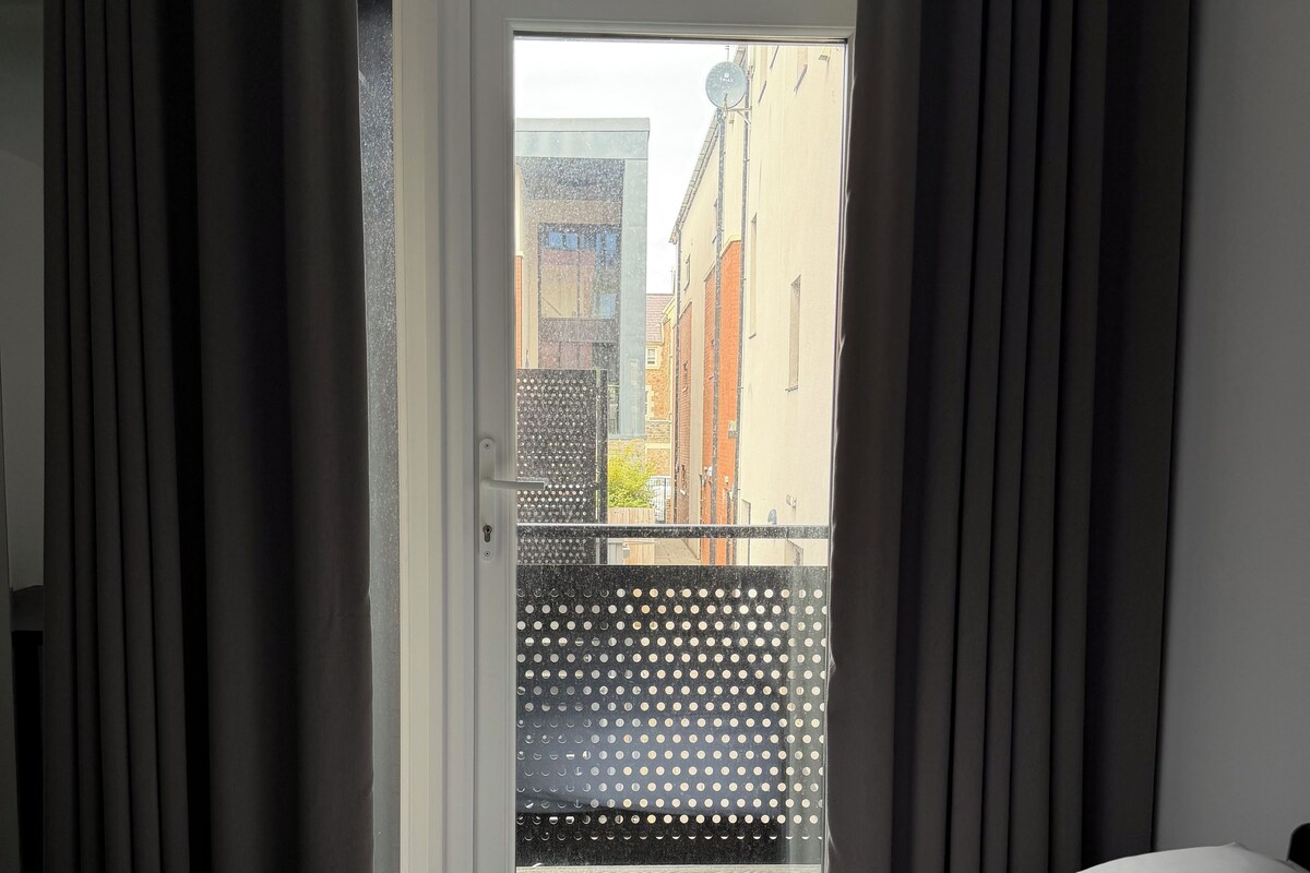 A hotel room with a bed, a window with curtains, and a view of buildings outside.