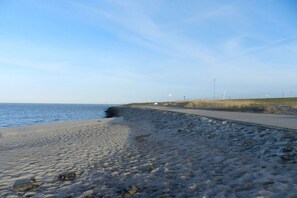 A sandy beach with a paved walkway, a grassy area, and wind turbines in the distance. - Holiday home with 4 bedrooms (Hooksiel Wangerland)