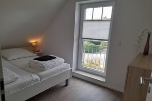 A bedroom with a bed, a bedside lamp, a window with blinds, and a wooden cabinet. - Holiday home with 4 bedrooms (Hooksiel Wangerland)