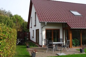 A house with a red-tiled roof, a patio with a table and chairs, and a garden with a hedge and a bicycle. - Holiday home with 3 bedrooms (Boltenhagen)