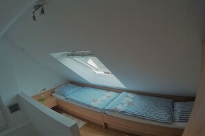 Ein kompakter Raum mit schräger Decke, einem Oberlicht, einem Bett mit gestreifter Bettwäsche, einem Nachttisch mit Lampe und einem kleinen Fenster.