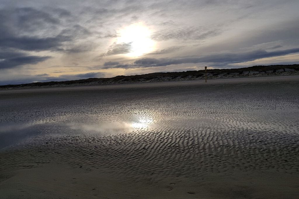 A beach with a wet sand surface reflecting the sky, a distant dune, and a solitary structure.