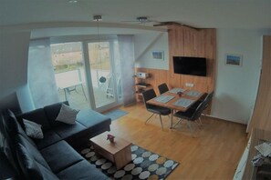 A modern living room with a sofa, dining table, and chairs. There is a TV mounted on the wall, and a sliding glass door leading to an outdoor area. - Holiday apartment with 2 bedrooms (Norderney)