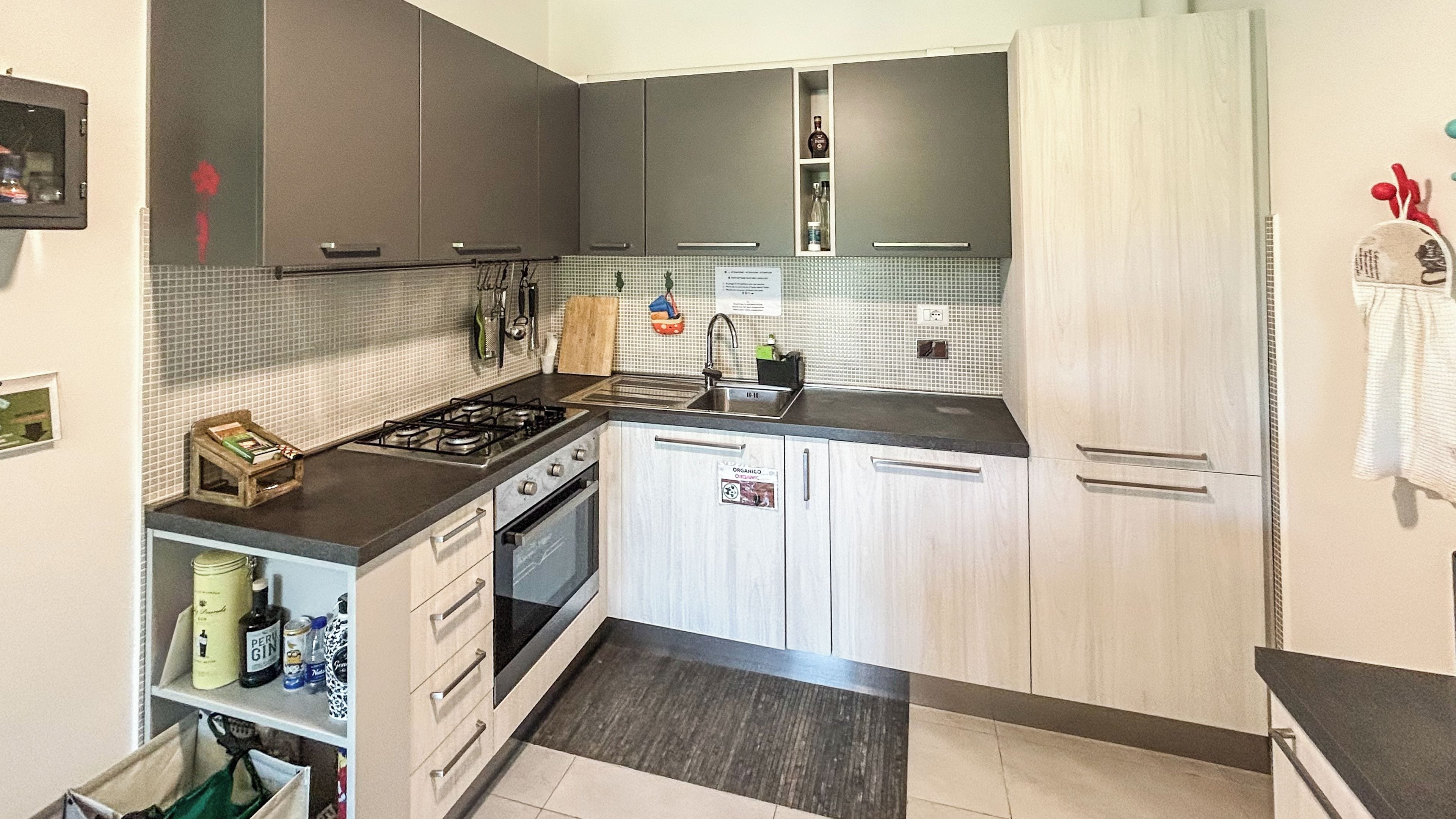 Una cucina compatta con piano cottura a gas, forno e lavello. Si vede uno scaffale con bottiglie e una borsa verde.