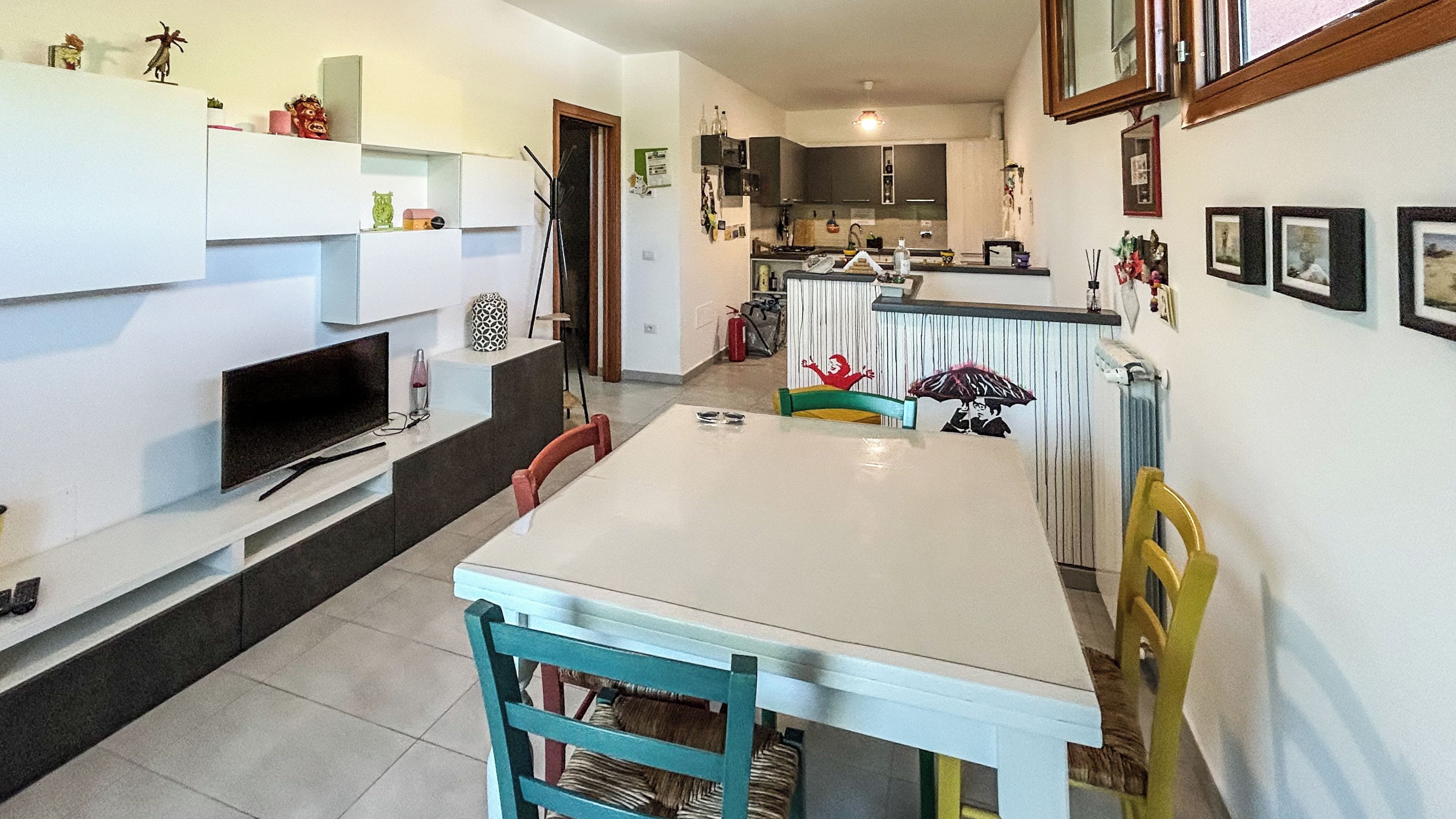 Un coin repas avec une table et des chaises blanches, un téléviseur posé sur un meuble, et des meubles de cuisine en arrière-plan.