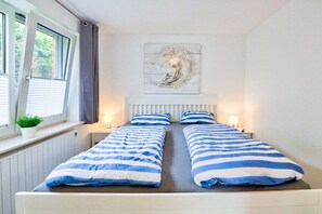 A double bed with striped bedding, a window with curtains, a radiator, and a wall-mounted artwork. - Fasanenweg Holiday Apartment 2 (St. Peter-Ording)