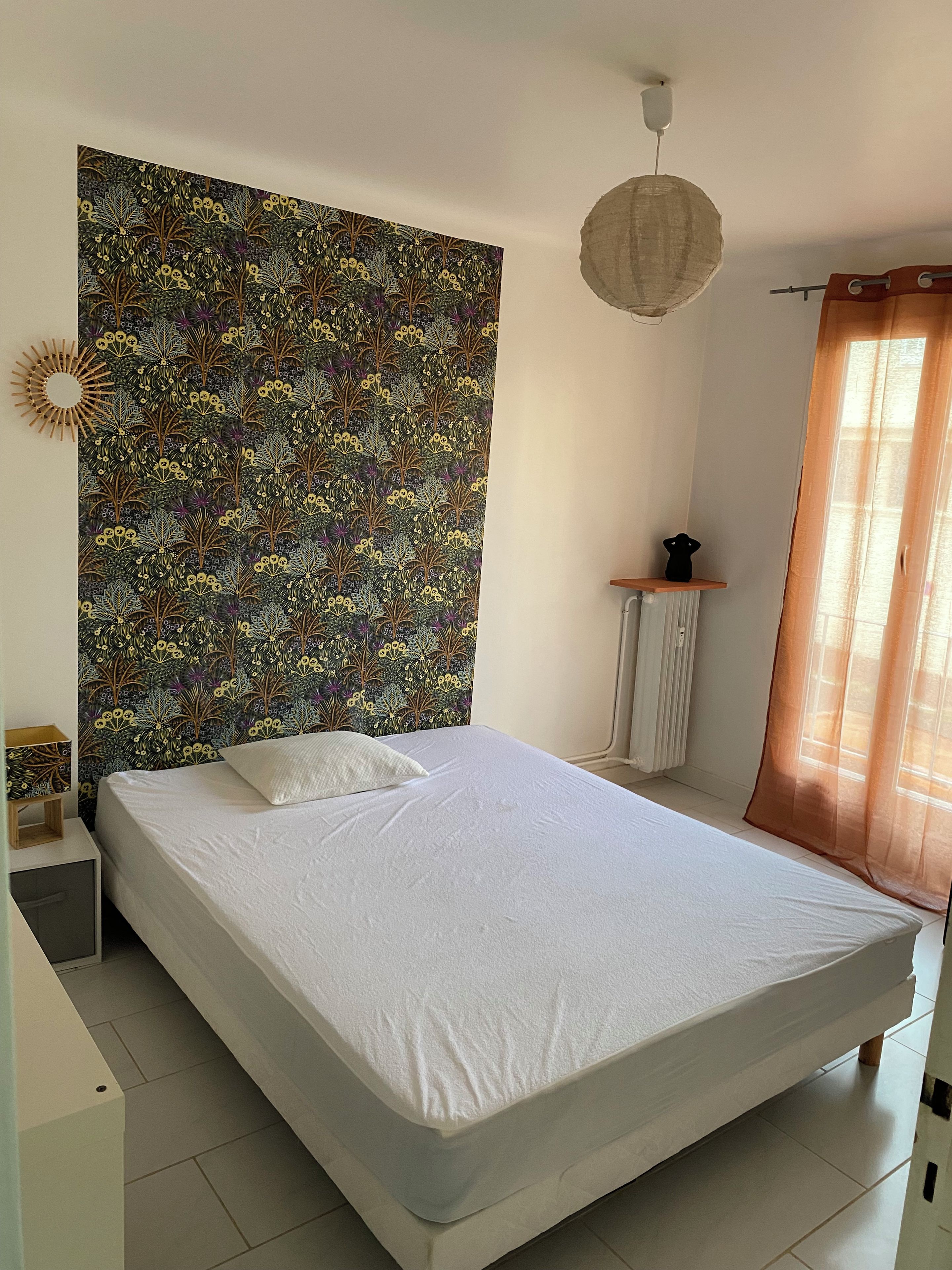 A bedroom with a bed, a headboard with a floral pattern, a bedside table, a lamp, and a window with curtains.