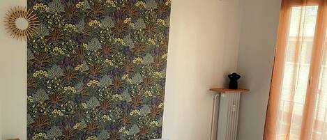 A bedroom with a bed, a headboard with a floral pattern, a bedside table, a lamp, and a window with curtains.