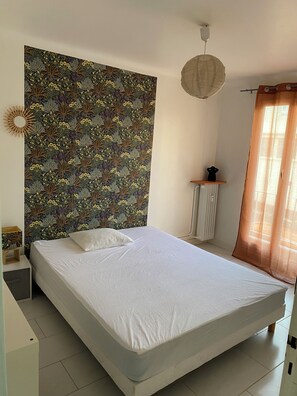 Ein Schlafzimmer mit einem Bett, einem Kopfschild mit Blumenmuster, einem Nachttisch, einer Lampe und einem Fenster mit Vorhängen.